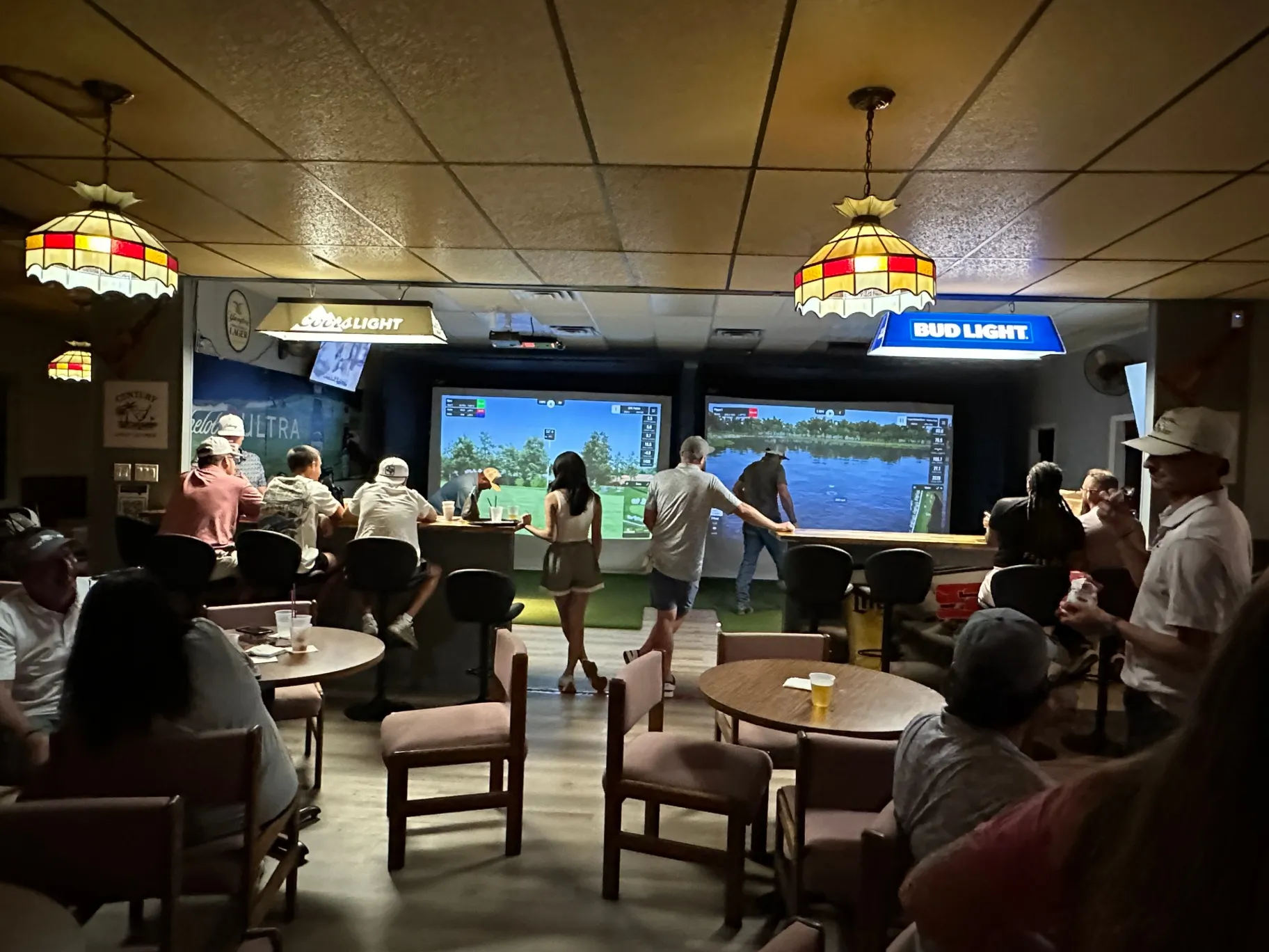 Group of people enjoying golf simulations in a lively bar setting with vintage lamps and beer advertisements Group of people enjoying golf simulations in a lively bar setting with vintage lamps and beer advertisements