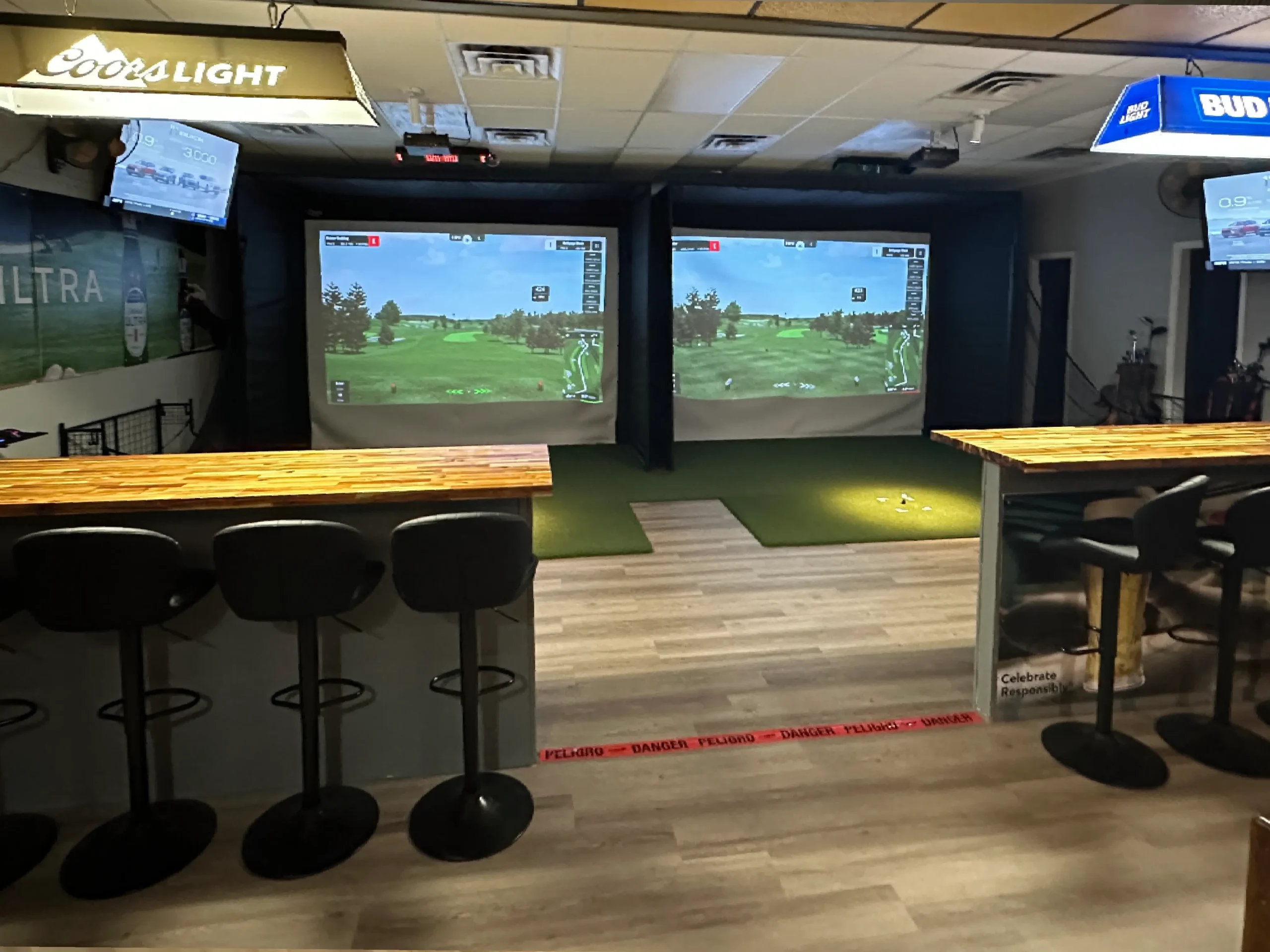 Indoor golfing facility featuring two projection screens, a wooden bar, and seating, with screens displaying golf courses and monitors (1) Indoor golfing facility featuring two projection screens, a wooden bar, and seating, with screens displaying golf courses and monitors
